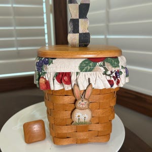 May include: A decorative Easter basket with a wooden lid and a fabric trim featuring a fruit pattern. The basket has a wooden bunny accent and a separate wooden square. The basket is made of woven material.