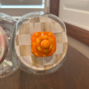 May include: A clear glass jar lid featuring a beige and white checkered pattern. Centered on the lid is a bright orange knob with a spiral design and yellow polka dots. The knob has a tiered, circular shape that rises from the center of the lid.