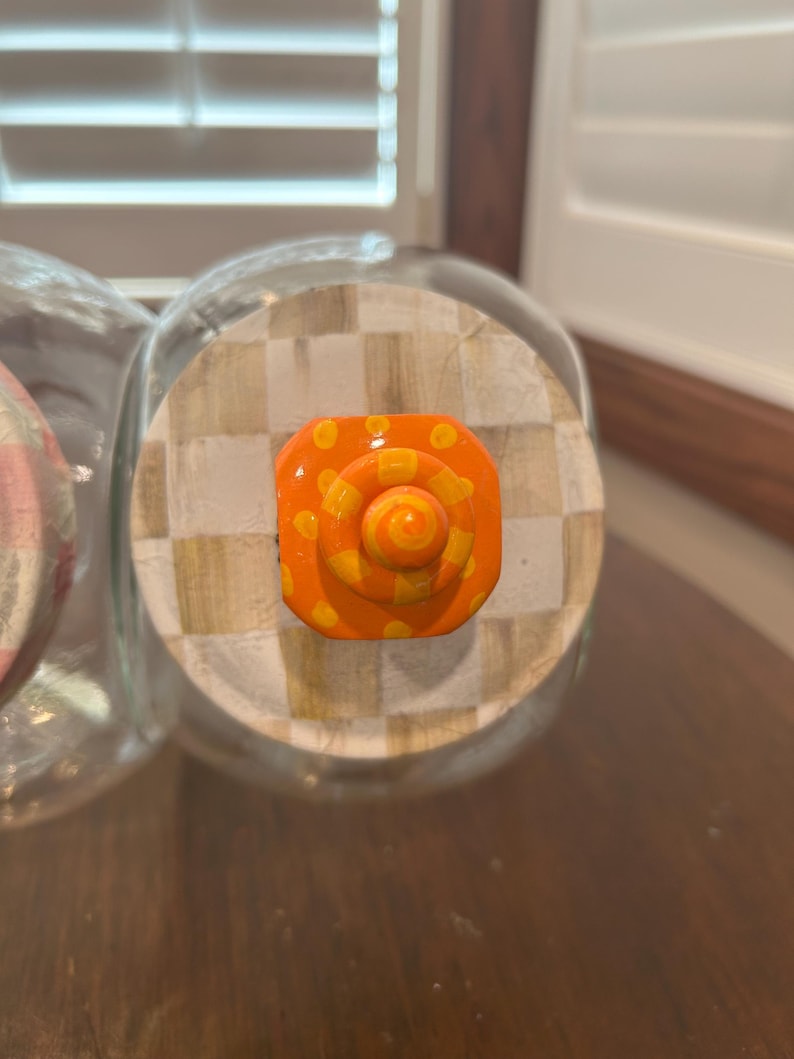 May include: A clear glass jar lid featuring a beige and white checkered pattern. Centered on the lid is a bright orange knob with a spiral design and yellow polka dots. The knob has a tiered, circular shape that rises from the center of the lid.