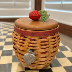 May include: A round woven basket with a natural tan and red splint pattern, featuring a wooden lid topped with a red strawberry and green leaf. A silver-toned metal strawberry charm hangs from the rim, which is accented by a red band with nails.