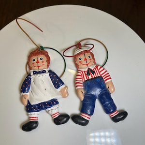 May include: Two Raggedy Ann and Andy doll ornaments. The Raggedy Ann doll is wearing a blue dress with white apron. The Raggedy Andy doll is wearing a red and white striped shirt and blue overalls. Both have black shoes and red yarn hair.
