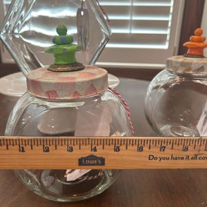 May include: Two clear glass jars with round bodies and lids decorated in pink and white checkered patterns. The front jar features a green and purple finial and is measured by a wooden ruler showing a diameter of approximately 5 inches.