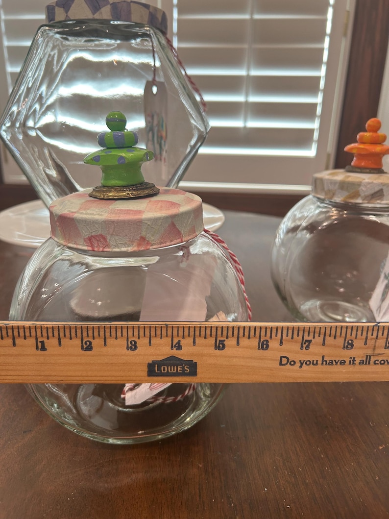 May include: Two clear glass jars with round bodies and lids decorated in pink and white checkered patterns. The front jar features a green and purple finial and is measured by a wooden ruler showing a diameter of approximately 5 inches.