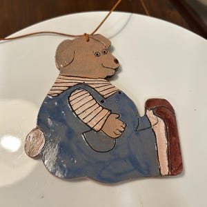 May include: A ceramic ornament of a brown bear wearing blue overalls and a striped shirt. The bear is holding a red and brown object in its right paw.