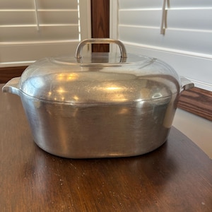 May include: A vintage, silver-toned metal casserole dish with a lid and a handle. The dish has a rounded shape with side handles and sits on a dark wooden surface. The background includes white shutters and a wooden frame.