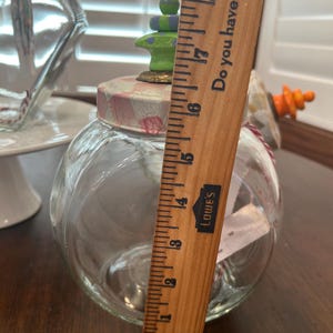 May include: A clear glass jar with a round body and a lid featuring a pink and white checkered pattern. A green and purple stacked finial sits on top. A wooden ruler placed vertically shows the jar stands approximately 6.5 inches tall.