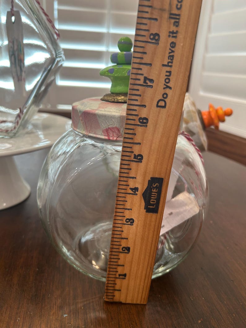 May include: A clear glass jar with a round body and a lid featuring a pink and white checkered pattern. A green and purple stacked finial sits on top. A wooden ruler placed vertically shows the jar stands approximately 6.5 inches tall.