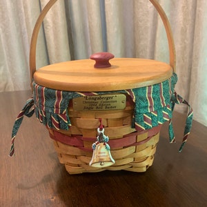 May include: A woven basket with a wooden lid and handle. The basket has a green and red striped fabric trim and a decorative bell charm. The lid has a small, round, maroon knob. The basket is labeled "Longaberger Christmas Collection 1994 Edition Jingle Bell Basket."