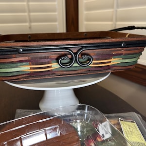 May include: A rectangular woven basket with a dark brown wooden frame and black metal handles. The basket features a mix of colorful woven strips in shades of green, purple, and orange. It sits on a white cake stand.