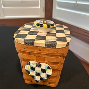 May include: A square woven wooden basket with a lid featuring a black and white checkerboard pattern. A ceramic knob with a matching checkerboard design sits on top. A heart-shaped wooden tag with a painted black, white, and blue check pattern hangs from the front.