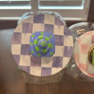 May include: A circular jar lid featuring a hand-painted purple and white checkerboard pattern. Centered on the lid is a square, light purple knob decorated with bright lime green polka dots and a central lime green spiral design.