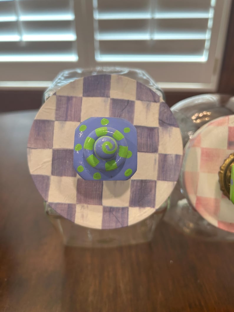 May include: A circular jar lid featuring a hand-painted purple and white checkerboard pattern. Centered on the lid is a square, light purple knob decorated with bright lime green polka dots and a central lime green spiral design.