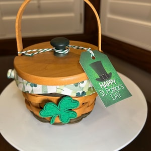 May include: A wooden basket with a handle and lid, featuring a green felt shamrock and a tag that reads "Happy St. Patrick's Day." The basket has a green and white patterned fabric trim.