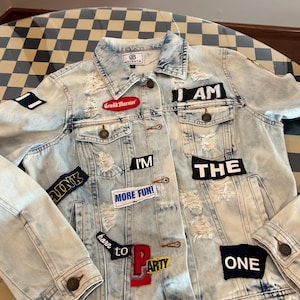 May include: A light-wash denim jacket featuring distressed areas and various embroidered patches. The patches include text such as I AM, THE, ONE, I'M, MORE FUN, and BORN TO PARTY, along with a red Grand Marnier logo and a small blue rectangular patch.