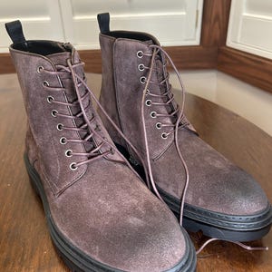May include: A pair of brown suede boots with a lace-up design and black soles. The boots have a side zipper and a pull tab at the top. The boots are sitting on a wooden surface.