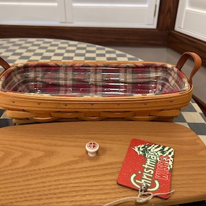 May include: A rectangular woven wood basket with two leather handles and a plaid fabric liner in red, green, and tan. A matching wooden lid with a small knob sits in front, accompanied by a red tag reading Christmas WISHES.
