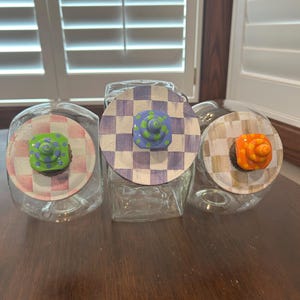 May include: Three clear glass canisters with round lids. The left lid has a pink and white checkered pattern with a green knob. The center lid has a purple and white checkered pattern with a blue knob. The right lid has a tan and white checkered pattern with an orange knob.