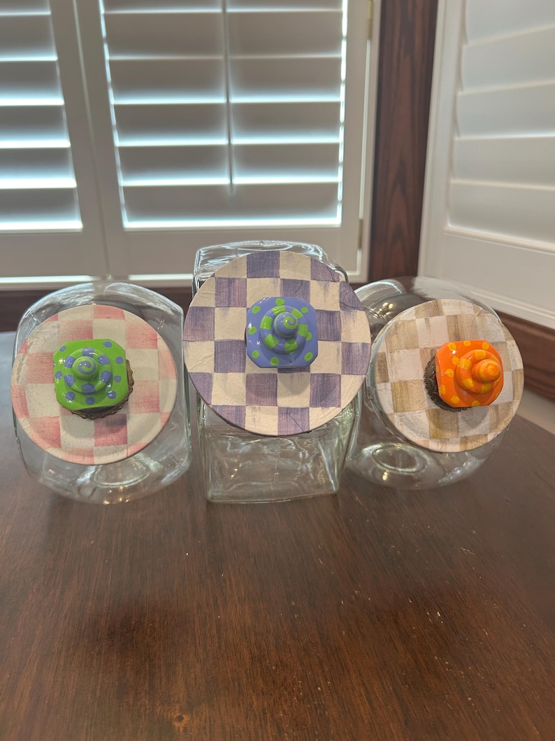 May include: Three clear glass canisters with round lids. The left lid has a pink and white checkered pattern with a green knob. The center lid has a purple and white checkered pattern with a blue knob. The right lid has a tan and white checkered pattern with an orange knob.
