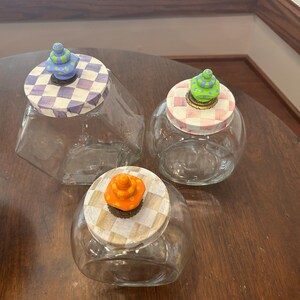 May include: Three clear glass jars with decorative lids. One lid has a purple and white checkerboard pattern with a blue and green finial. Another has a pink and white checkerboard pattern with a green finial. The third has a tan and white checkerboard pattern with an orange finial.