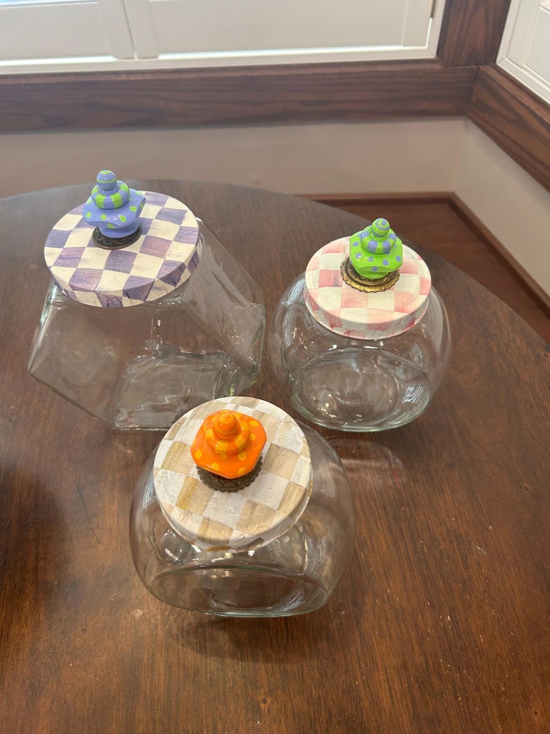 May include: Three clear glass jars with decorative lids. One lid has a purple and white checkerboard pattern with a blue and green finial. Another has a pink and white checkerboard pattern with a green finial. The third has a tan and white checkerboard pattern with an orange finial.