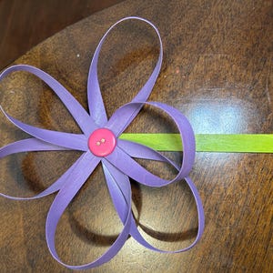 May include: A handmade paper flower with lavender petals and a pink button center. The flower is attached to a green stem. The craft is set against a wooden surface.