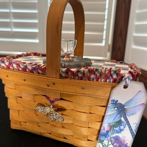 May include: A woven light brown wooden basket with a single arched handle. The lid features a blue and white checkered fabric with a ruffled red and white trim. A clear glass knob sits on top. A gold bee charm and a dragonfly-themed tag are attached.