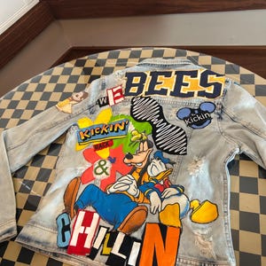 May include: A light-wash denim jacket featuring various colorful patches and embroidery on the back. The design includes the text "BEES", "KICKIN BACK & CHILLIN", and "Kickin", along with illustrations of Goofy and Donald Duck, plus a black wave pattern.