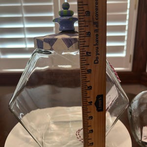 May include: A clear glass hexagonal jar with a decorative lid featuring a purple and white checkered pattern and a blue and green finial. A wooden ruler placed against the jar indicates a height of approximately 10 inches from base to top.