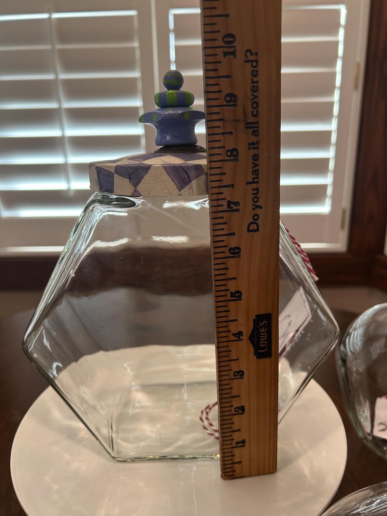 May include: A clear glass hexagonal jar with a decorative lid featuring a purple and white checkered pattern and a blue and green finial. A wooden ruler placed against the jar indicates a height of approximately 10 inches from base to top.