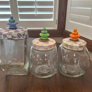 May include: Three clear glass canisters with lids featuring checkered patterns and decorative finials. The left lid has a blue and white check with a blue and green finial. The middle lid has a pink and white check with a green finial. The right lid has a tan and white check with an orange finial.