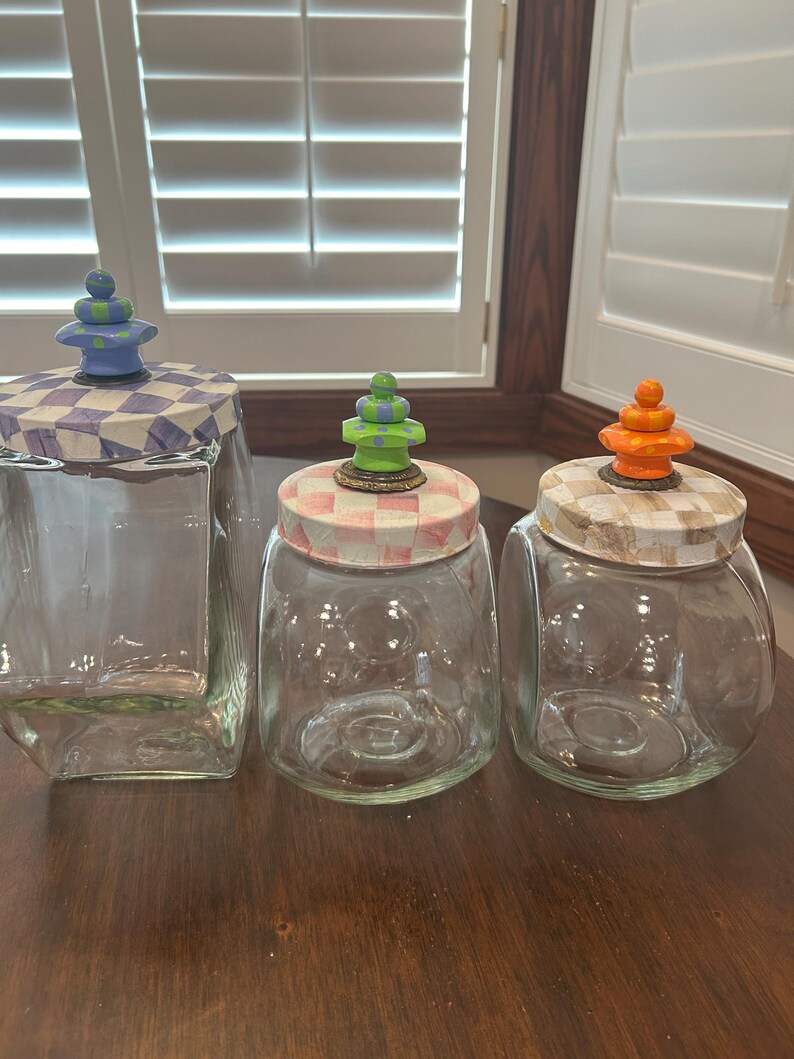 May include: Three clear glass canisters with lids featuring checkered patterns and decorative finials. The left lid has a blue and white check with a blue and green finial. The middle lid has a pink and white check with a green finial. The right lid has a tan and white check with an orange finial.