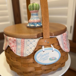 May include: A brown woven basket with a wooden lid and handle. The basket has a decorative fabric lining with pink and blue patterns. A ceramic Easter-themed ornament is attached, reading "Happy Easter 2003."