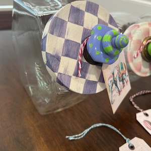 May include: A clear glass jar with a circular lid featuring a purple and white checkered pattern. The lid is topped with a blue and green polka-dot knob and a red and white twine string. A rectangular tag with an elephant illustration hangs from the lid.