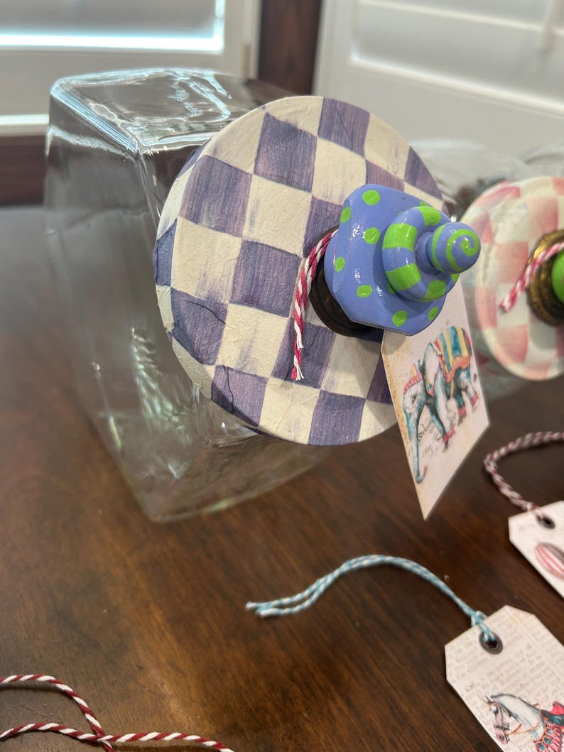 May include: A clear glass jar with a circular lid featuring a purple and white checkered pattern. The lid is topped with a blue and green polka-dot knob and a red and white twine string. A rectangular tag with an elephant illustration hangs from the lid.