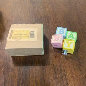 May include: A brown cardboard box with a label that reads "Longaberger" and "Knob - Birthday Baby" next to four colorful toy blocks spelling "BABY". The blocks are blue, pink, green, and yellow.