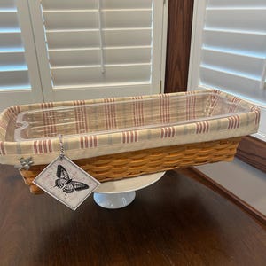May include: A rectangular woven basket with a red and cream striped fabric lining and a clear plastic insert. A tag with a butterfly illustration hangs from the basket. The basket is on a white cake stand.