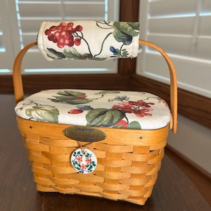 May include: A Longaberger basket with a lid and handle. The basket is made of woven light brown wood. The lid and handle are covered in a fabric with a floral pattern of red grapes and green leaves. A small decorative tag hangs from the front.
