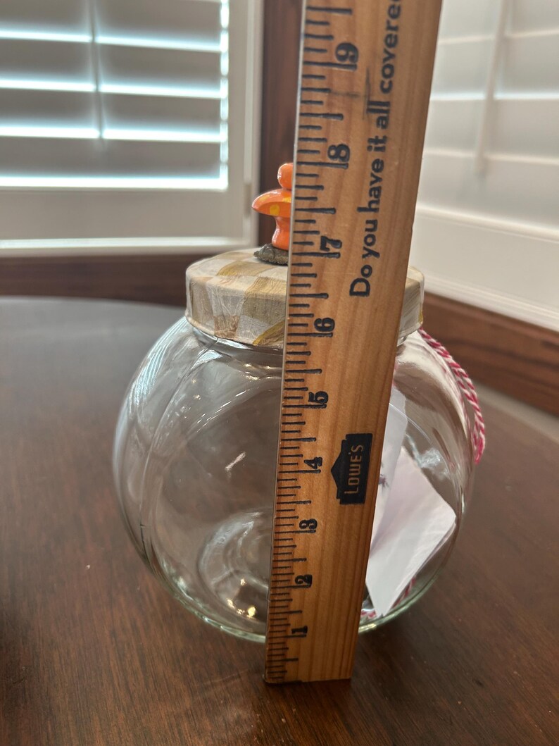 May include: A clear glass jar with a pumpkin-shaped body and a lid topped with a small orange mushroom figure. A wooden ruler is placed vertically against the jar, showing a height measurement of approximately 7 inches from the base to the lid.