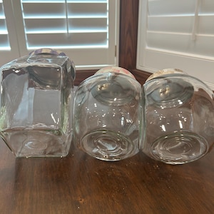May include: Three clear glass storage jars with lids. One jar is rectangular with a purple and white checkered lid. Two jars are rounded with pink and white checkered lids. The jars are empty and feature circular indentations on their bases.
