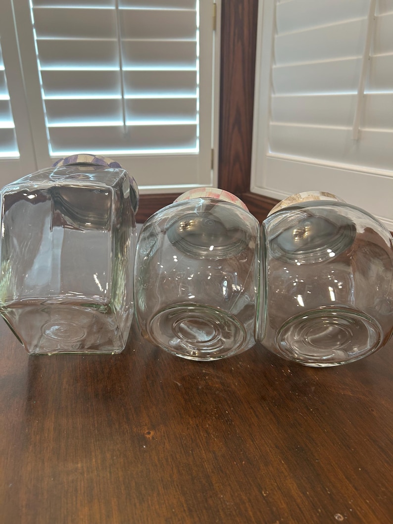 May include: Three clear glass storage jars with lids. One jar is rectangular with a purple and white checkered lid. Two jars are rounded with pink and white checkered lids. The jars are empty and feature circular indentations on their bases.