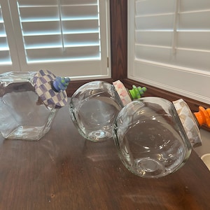 May include: Three clear glass jars with angled openings. Each jar features a unique decorative lid: one with a blue and white checkered pattern and blue knob, one with a pink floral pattern and green knob, and one with a tan pattern and orange knob.