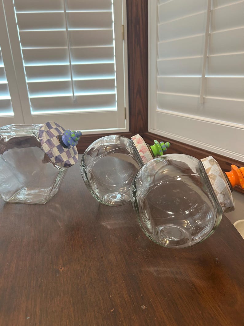 May include: Three clear glass jars with angled openings. Each jar features a unique decorative lid: one with a blue and white checkered pattern and blue knob, one with a pink floral pattern and green knob, and one with a tan pattern and orange knob.