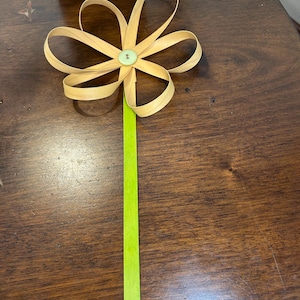May include: A handmade wooden flower with six looped petals and a green button center. The flower sits atop a long, bright green stem. The flower is made of thin, light-colored wood strips.