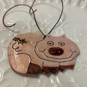 May include: A hand-painted ceramic ornament shaped like a pig with a brown and pink glaze. The pig has a smiling face with a brown snout and eyes. The ornament has a gold star on its back and a wire loop for hanging.