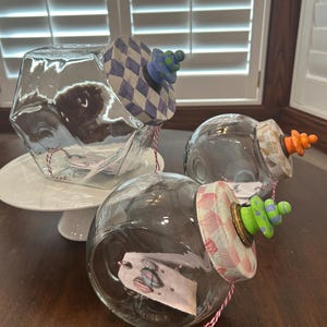 May include: Three clear glass jars with angled openings, each featuring a decorative lid with a checkered pattern and a colorful stacked bead finial. The lids are painted in blue, orange, and pink, with matching red and white twine tags inside.