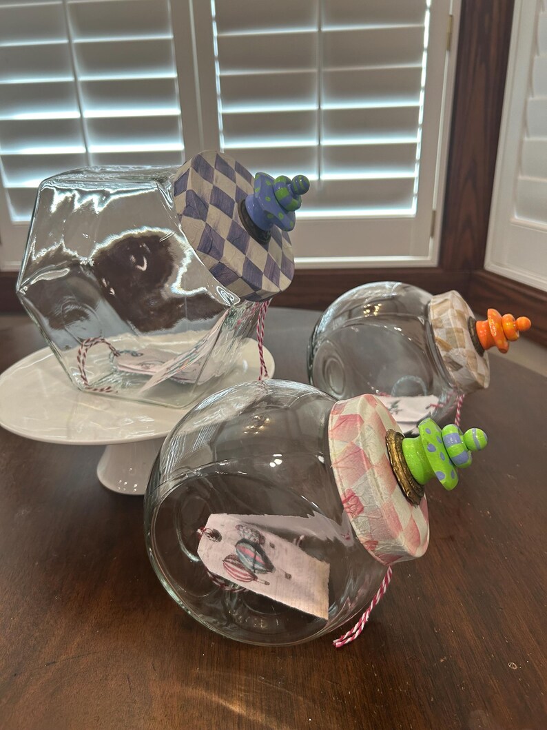 May include: Three clear glass jars with angled openings, each featuring a decorative lid with a checkered pattern and a colorful stacked bead finial. The lids are painted in blue, orange, and pink, with matching red and white twine tags inside.