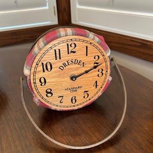 May include: A round wooden clock with the word "DRESDEN" and "STANDARD TIME" printed on the face. The clock has black numerals and hands, and is surrounded by a red and tan plaid fabric. The clock is supported by a curved metal stand.