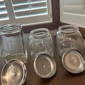 May include: Three clear glass storage jars with wide openings and matching metal lids. The lids feature a decorative pattern on the exterior and a silver-toned interior with a central screw. The jars have a square base and rounded corners.