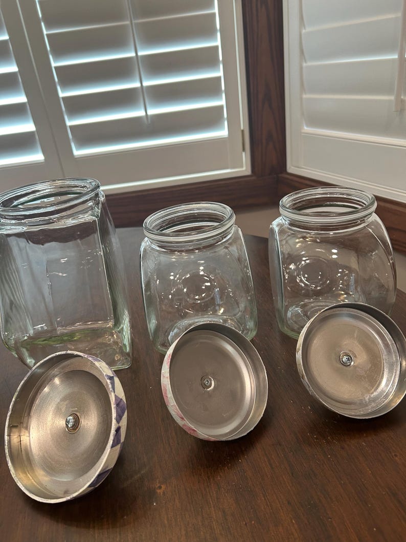 May include: Three clear glass storage jars with wide openings and matching metal lids. The lids feature a decorative pattern on the exterior and a silver-toned interior with a central screw. The jars have a square base and rounded corners.