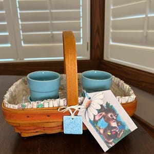 May include: A woven basket with a wooden handle, holding two small blue pots. The basket has a floral patterned lining and a tag with a bunny illustration. A blue square tag with a flower design is attached.